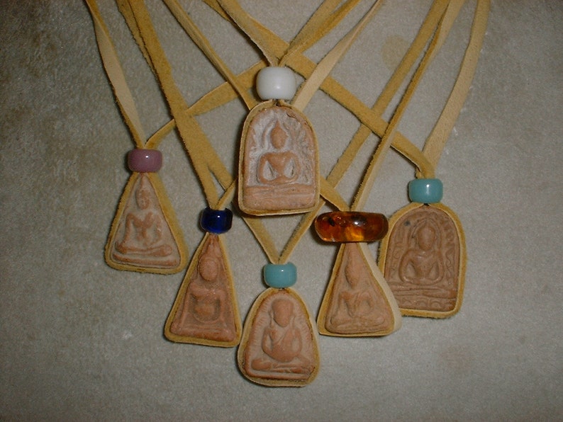 K&ouml;nnte beinhalten: Sieben kleine, braune, tonfarbene Anh&auml;nger mit Bildern von Buddha, die jeweils an einer d&uuml;nnen, hellbraunen Lederschnur mit einer kleinen, bunten Glasperle h&auml;ngen.