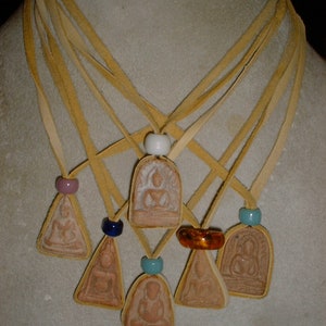 K&ouml;nnte beinhalten: Sieben Anh&auml;nger mit Bildern von Buddha, jeder an einer separaten Schnur aus braunem Leder. Die Schn&uuml;re sind oben mit einer wei&szlig;en Perle zusammengebunden. Die Anh&auml;nger sind alle in verschiedenen Formen und Gr&ouml;&szlig;en, und einige haben farbige Perlen.
