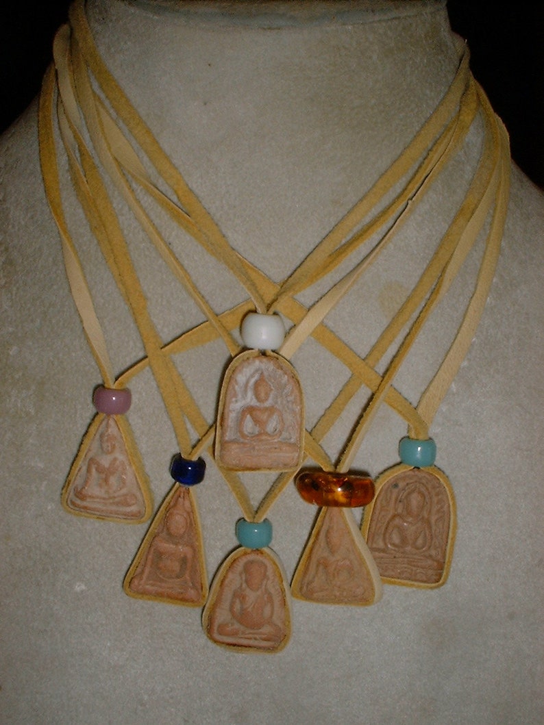 K&ouml;nnte beinhalten: Sieben Anh&auml;nger mit Bildern von Buddha, jeder an einer separaten Schnur aus braunem Leder. Die Schn&uuml;re sind oben mit einer wei&szlig;en Perle zusammengebunden. Die Anh&auml;nger sind alle in verschiedenen Formen und Gr&ouml;&szlig;en, und einige haben farbige Perlen.