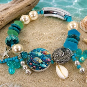 May include: A beaded bracelet with a nautical theme. The bracelet features a variety of beads in shades of blue, green, and cream, including pearls, turquoise-colored stones, and a round charm with a fish design. Additional charms include a starfish, a seashell, and a silver-colored spiral.