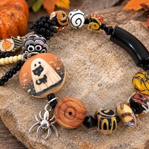 Könnte beinhalten: Ein Perlenarmband mit einem großen runden Anhänger, der ein Gespenst mit einer schwarzen Katze zeigt, einem silbernen Spinnenanhänger sowie verschiedenen Perlen in Schwarz, Orange, Weiß und Gelb. Das Armband enthält gemusterte Glasperlen, eine schwarze Röhrenperle und Holzakzente.