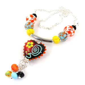 Könnte beinhalten: Eine farbenfrohe Halskette mit einem herzförmigen Anhänger mit floralem Design und Wirbel. Die Halskette umfasst eine Vielzahl von Perlen in Rot, Orange, Gelb, Blau und Weiß, auf einer Silberkette aufgereiht.