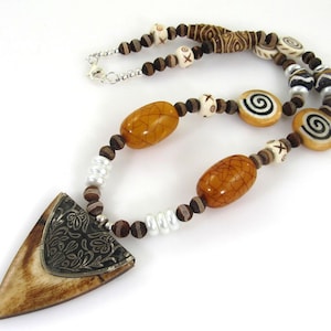 May include: A brown, white, and black beaded necklace with a large, triangular pendant. The pendant is made of bone and has a silver design. The necklace is made of various beads, including wood, bone, and glass.
