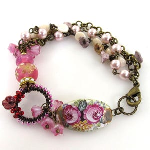 以下が含まれることがあります： ピンクと白のビーズ、花柄のセラミックビーズ、真鍮製の留め具が付いたマルチストランドブレスレットです。