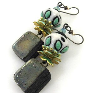 以下が含まれることがあります： 正方形のセラミックペンダントと緑色のアクセントが付いた白いセラミックビーズが付いた、ぶら下がりのイヤリングのペア。ビーズは金色の花のデザインで飾られています。