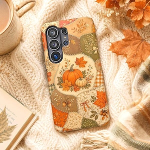 May include: A smartphone case featuring a patchwork design of autumn-themed illustrations, including orange pumpkins, brown acorns, and vibrant maple leaves in shades of gold, rust, and green, with a multi-lens camera cutout on the upper left side.