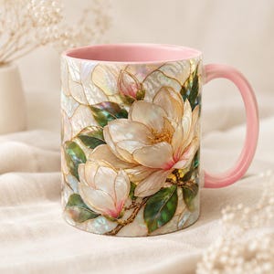 May include: A ceramic mug featuring a pearlescent, stained-glass style design of white magnolia flowers with green leaves and pink accents. The mug has a solid light pink handle and a matching light pink interior, showcasing a smooth finish.
