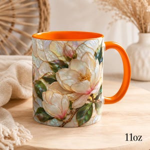 May include: A ceramic mug with an 11oz capacity, featuring a vibrant orange interior and handle. The exterior displays a detailed, iridescent floral pattern of white magnolia blossoms with green leaves, rendered in a stained-glass style.