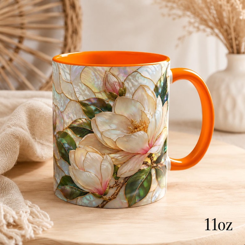 May include: A ceramic mug with an 11oz capacity, featuring a vibrant orange interior and handle. The exterior displays a detailed, iridescent floral pattern of white magnolia blossoms with green leaves, rendered in a stained-glass style.