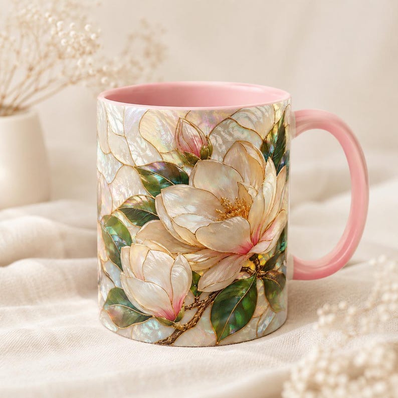 May include: A ceramic mug featuring a pearlescent, stained-glass style design of white magnolia flowers with green leaves and pink accents. The mug has a solid light pink handle and a matching light pink interior, showcasing a smooth finish.