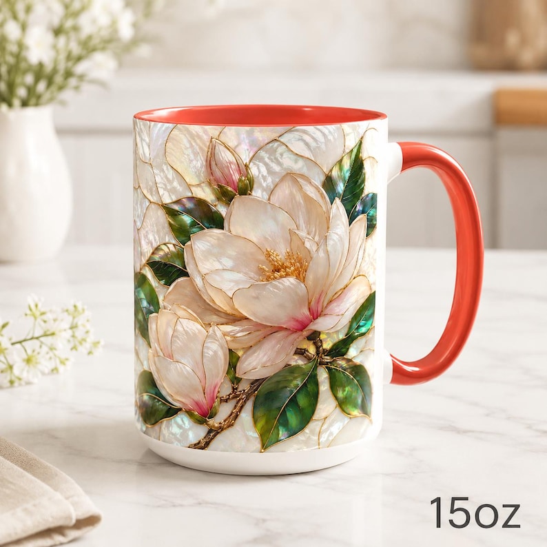 May include: A 15 oz ceramic mug featuring a stained glass style illustration of white magnolia flowers with pink accents and green leaves. The mug has a bright orange interior and a matching orange handle, with a white exterior base.