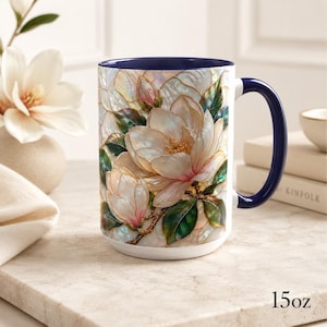 May include: A 15oz ceramic mug featuring a vibrant, iridescent-style illustration of white magnolia flowers with green leaves. The mug has a deep navy blue interior and a matching navy blue handle, with the text 15oz printed on the surface.