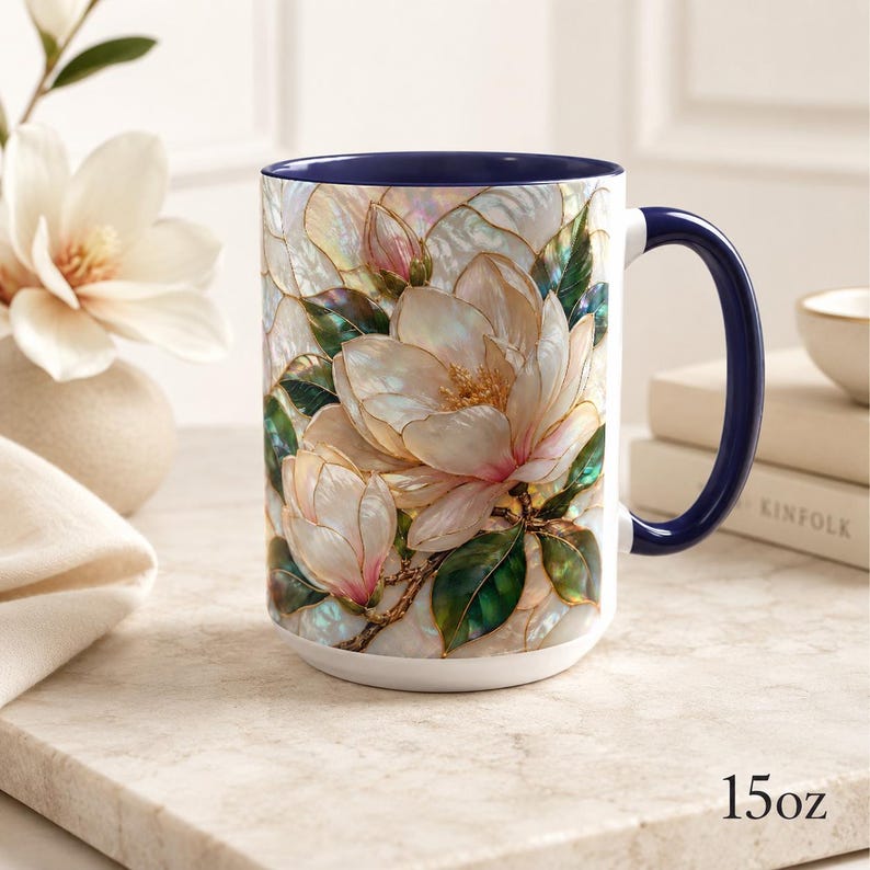 May include: A 15oz ceramic mug featuring a vibrant, iridescent-style illustration of white magnolia flowers with green leaves. The mug has a deep navy blue interior and a matching navy blue handle, with the text 15oz printed on the surface.