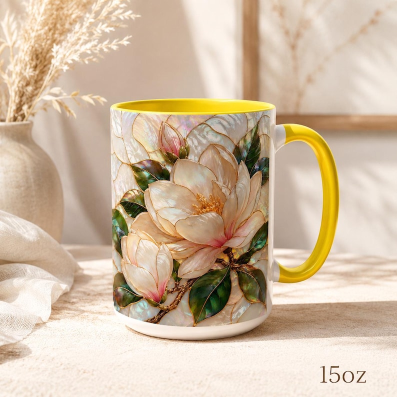May include: A 15oz ceramic mug featuring a vibrant yellow interior and handle. The exterior displays a detailed illustration of white and pink magnolia flowers with green leaves, rendered in a stained-glass style with iridescent, pearlescent accents.