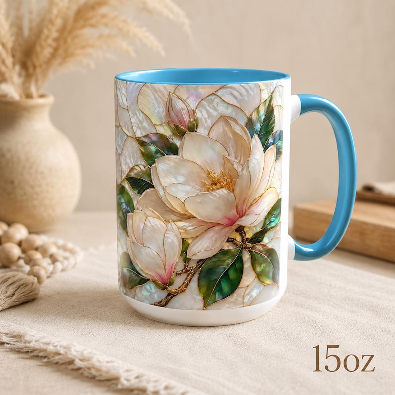 May include: A 15oz ceramic mug featuring a vibrant, stained-glass style illustration of white magnolia flowers with green leaves and gold outlines. The mug has a bright blue interior and a matching blue handle, set against a neutral background.