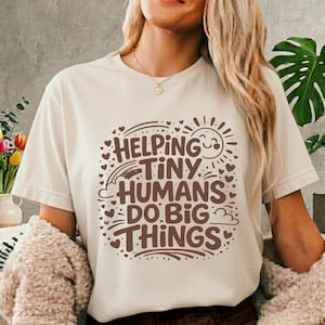 Puede incluir: Camiseta color crema con la frase marrón "Helping Tiny Humans Do Big Things". El diseño incluye un sol, un arcoíris, nubes y corazones. La camiseta tiene mangas cortas y cuello redondo.