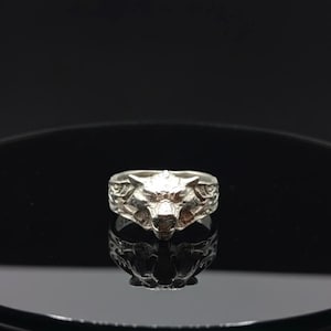 May include: A silver ring featuring a detailed wolf head design. The ring's band has an embossed pattern, and the wolf's face is prominently displayed. The ring is set against a dark, reflective surface.