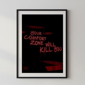Könnte beinhalten: Gerahmter Kunstdruck mit schwarzem Rand. Das Kunstwerk zeigt den Neon-Text "YOUR COMFORT ZONE WILL KILL YOU" in Rot vor einem dunklen Hintergrund. Der Druck wird an einer weißen Wand angezeigt.