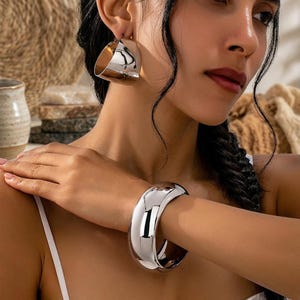 May include: A pair of polished silver-tone hoop earrings with a wide, curved design and a matching thick, smooth silver-tone cuff bracelet. Both pieces feature a high-shine metallic finish and a bold, rounded silhouette that catches the light.