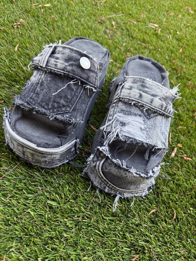 Peut inclure: Paire de sandales compens&eacute;es en jean d&eacute;lav&eacute; gris. Les sandales pr&eacute;sentent des bords effiloch&eacute;s, des boutons et une semelle &eacute;paisse. Elles sont pos&eacute;es sur de l'herbe verte.