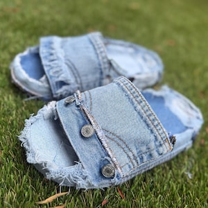 May include: A pair of light blue denim slide sandals with frayed edges and silver button details. The sandals are made from repurposed denim and sit on a green grassy surface.