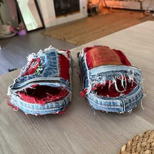 May include: A pair of open-toe slippers crafted from repurposed blue denim and red fabric. The slippers feature frayed edges, a visible zipper detail, and a tan leather Levi Strauss & Co. patch on the top. One slipper displays a floral patch.