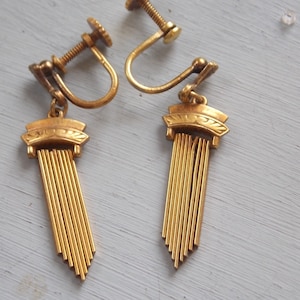 May include: A pair of gold tone clip-on earrings with a fringe design. The earrings have a decorative top with a floral pattern.