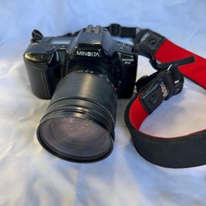 May include: Black Minolta Maxxum 5xi camera with a large lens and a red and black Aztec strap. The camera has the Minolta logo and lens measurements visible. The camera is on a white surface.