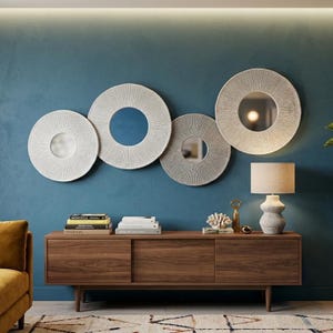 May include: Four round decorative wall mirrors with textured white frames and reflective centers are mounted on a teal wall. Below, a wooden cabinet with sliding doors holds books, a lamp, and decorative items.