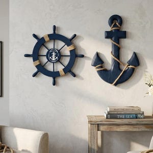 Op de afbeelding: Nautische wanddecoratie met een marineblauw scheepsstuur en anker. Het scheepsstuur heeft een wit embleem en is omwikkeld met touw. Het anker is ook marineblauw en omwikkeld met touw. De decoratie is op een lichtgekleurde muur gemonteerd.