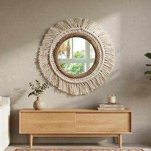 Peut inclure: Miroir rond en macramé beige avec bordure en corde et franges. Le miroir est accroché à un mur neutre. En dessous, un meuble en bois clair accueille un vase, de la verdure et une bougie. Le meuble mesure environ 120 cm de large.