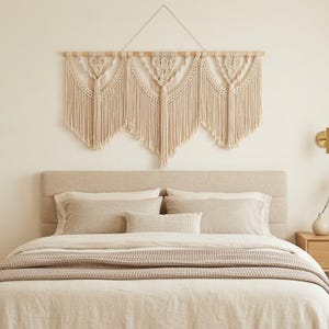 Könnte beinhalten: Ein beigefarbener Makramee-Wandbehang mit einer Holzstange und einer cremefarbenen Kordel. Das Makramee-Design weist geometrische Muster und Fransen auf und hängt über einem Bett mit neutralfarbener Bettwäsche.