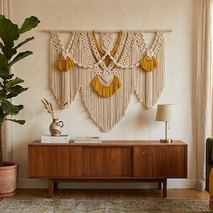Peut inclure: Une grande tenture murale en macramé composée de nœuds en corde de coton blanc cassé et de motifs tissés, agrémentée de sections de franges jaune moutarde vif. L'ensemble est suspendu à une tige en bois horizontale, créant un design superposé.