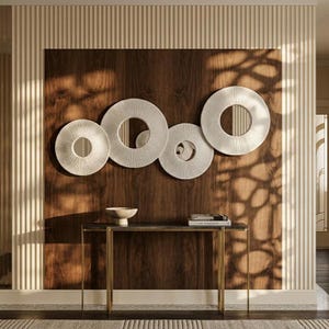 Peut inclure: Quatre éléments d'art mural circulaires blancs avec des surfaces texturées sont montés sur un panneau de bois foncé. En dessous, une table étroite avec un plateau en marbre et des pieds dorés supporte un petit bol et des livres. Le mur derrière la table a des panneaux verticaux crème.