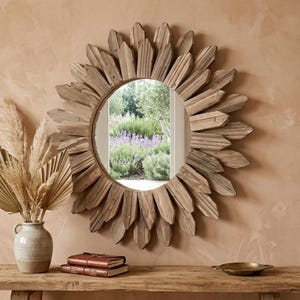 Peut inclure: Un miroir rond avec un cadre en forme de soleil en bois vieilli. Le miroir reflète une scène de jardin avec de la lavande et de la verdure. Il est fixé sur un mur pêche au-dessus d'une table en bois avec des objets décoratifs.