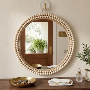 Peut inclure: Un miroir mural rond doté d'un cadre orné de deux rangées concentriques de perles en bois clair et d'une bordure en corde de jute épaisse. Le miroir est suspendu par une boucle en corde à un crochet mural transparent, reflétant une porte en bois.