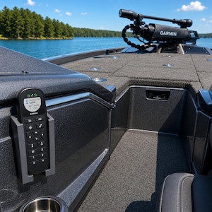 May include: A black Garmin remote control with a digital display is mounted on a gray boat console. The remote is in a gray holder, and the boat is on a lake. The display shows navigation data. A trolling motor is visible.