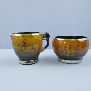 May include: A brown and black ceramic creamer and sugar bowl set with a silver rim. Both pieces feature a black and white illustration of a person riding a horse.