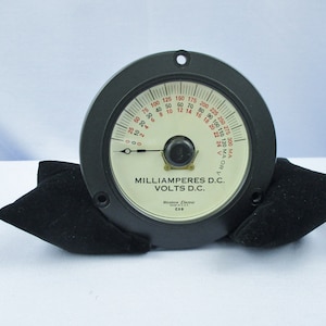 May include: A black circular analog meter with a white face and a black needle. The meter has a white dial with red markings and black text. The text on the dial reads "MILLIAMPERES D.C. VOLTS D.C.". The meter is made by "Western Electric" and has a serial number of "C98".