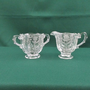May include: A clear glass sugar bowl and creamer set with a delicate etched floral design. The set is on a green background.