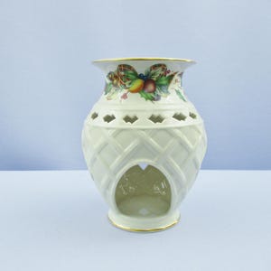 May include: Cream-colored ceramic vase with a lattice design and decorative top. The top features a gold rim and a festive pattern of bows, berries, and fruit. The vase has a large opening at the base.
