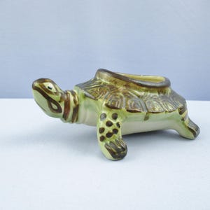 May include: A ceramic turtle figurine in shades of green, brown, and cream. The turtle has a hollowed-out back, suggesting it may be a planter or decorative item. The figurine is detailed with textured shell patterns and spotted legs.