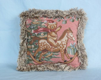 May include: Square decorative pillow with a Christmas theme. The pillow features a teddy bear riding a rocking horse in front of a Christmas tree. The pillow has a pink background and is trimmed with a fringed border.