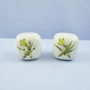 May include: A set of two white ceramic salt and pepper shakers with a floral design. The shakers have yellow flowers with green leaves and brown stems.