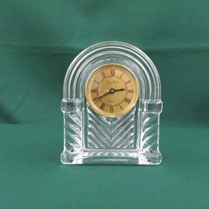 May include: A clear crystal clock with a gold face and Roman numeral hour markers. The clock is in the shape of an archway with a decorative design on the front. The clock face reads "Crystal Clear Quartz".