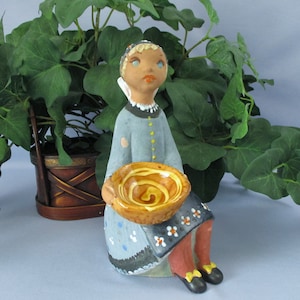 May include: A ceramic figurine of a girl wearing a blue dress and a white head scarf. She is holding a brown bowl with a yellow swirl design. The figurine is sitting on a light blue surface.