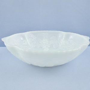 May include: A white, oval-shaped bowl with a scalloped rim. The bowl features a raised grape and leaf design. The bowl is made of opaque white glass and is set against a light blue background. This bowl is suitable for serving.
