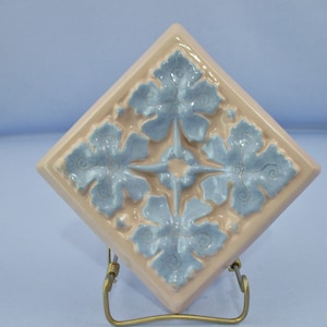 May include: A square ceramic tile with a raised design of four blue flowers on a pink background.