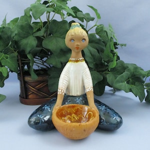 May include: A ceramic figurine of a girl sitting with a bowl in her lap. The girl has blonde hair, blue eyes, and is wearing a white shirt and blue pants with white flowers. The bowl is brown with a yellow interior.