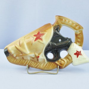 May include: A ceramic liquor bottle shaped like a gun with a red star on the side. The bottle is brown and black with a white handle. It is on a metal stand.